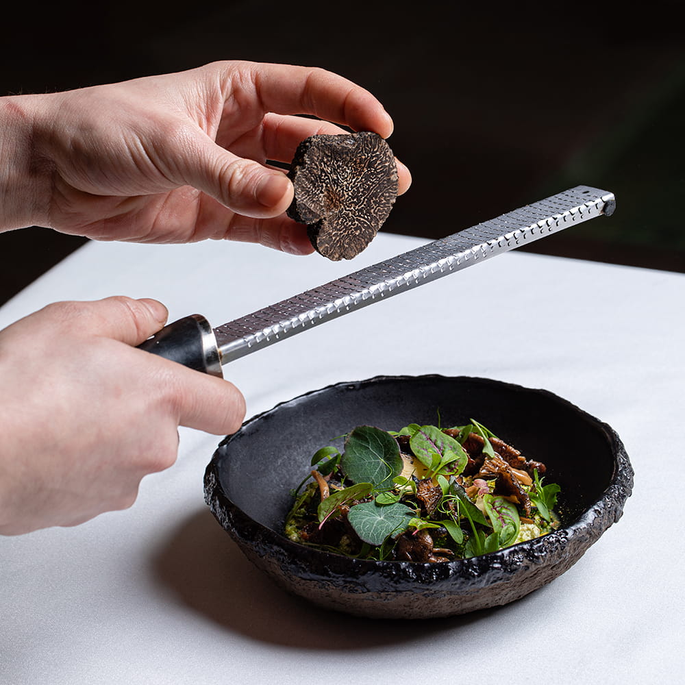Fotografía gastronómica de chef rallando trufa negra fresca sobre plato gourmet con vegetales y setas en restaurante