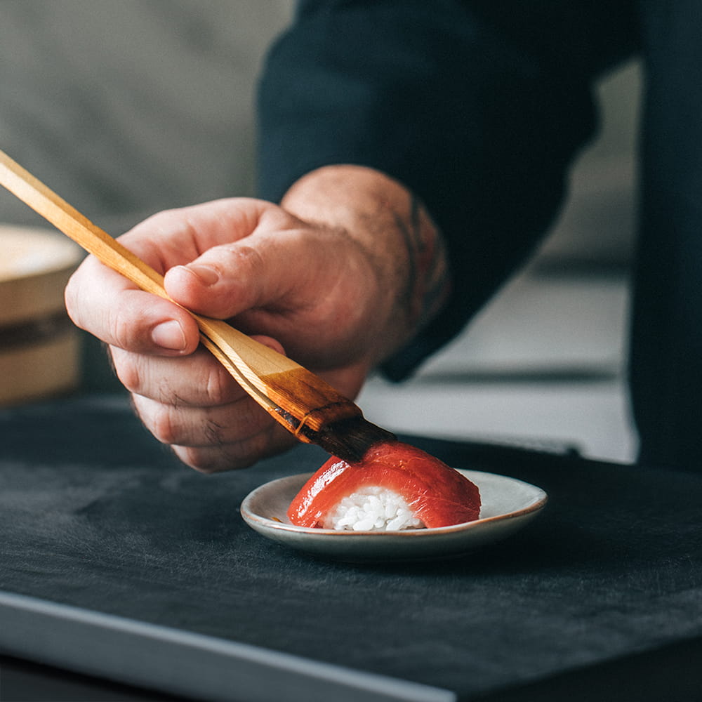 Fotografía gastronómica de chef pincelando nigiri de atún con salsa sobre arroz en cocina japonesa profesional