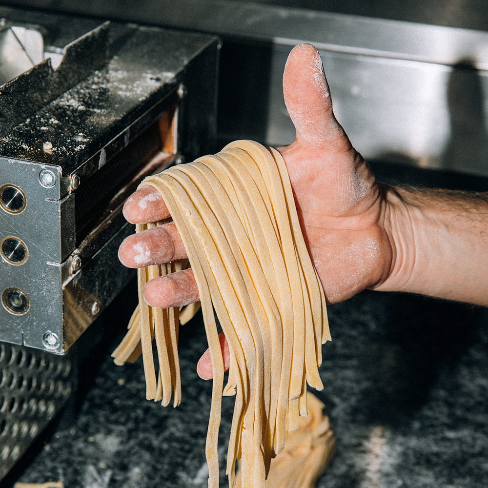 Fotografía gastronómica de pasta fresca recién cortada saliendo de máquina laminadora sobre manos de chef en cocina profesional