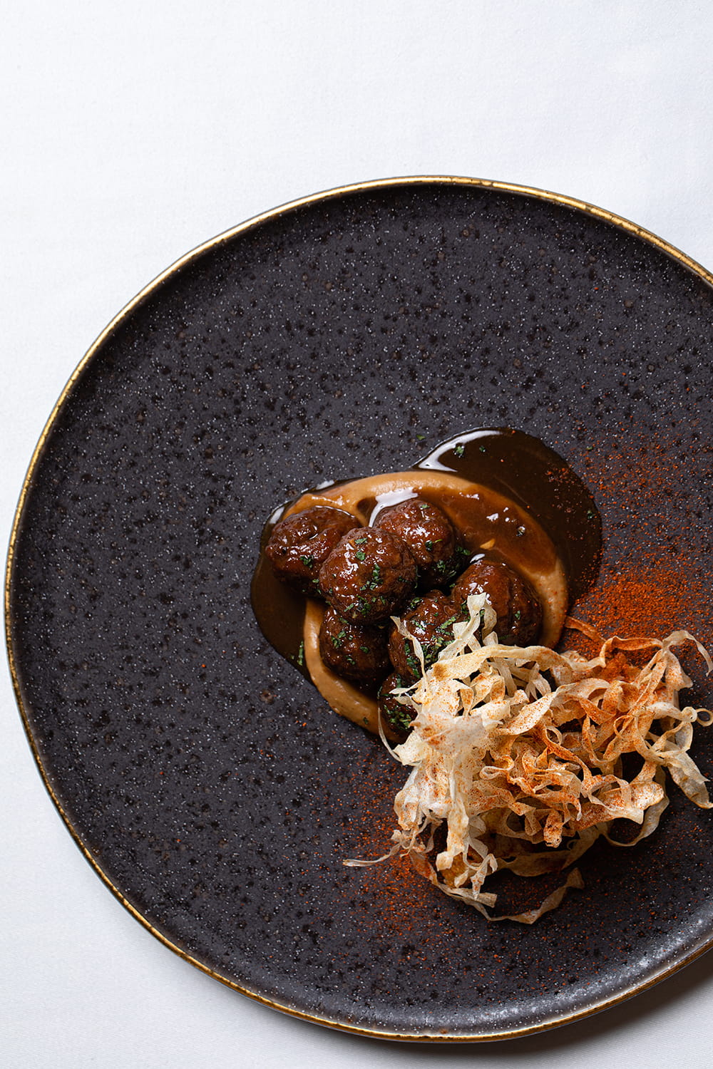 Fotografía gastronómica cenital de plato gourmet con albóndigas en salsa, crema y crujiente vegetal sobre vajilla negra