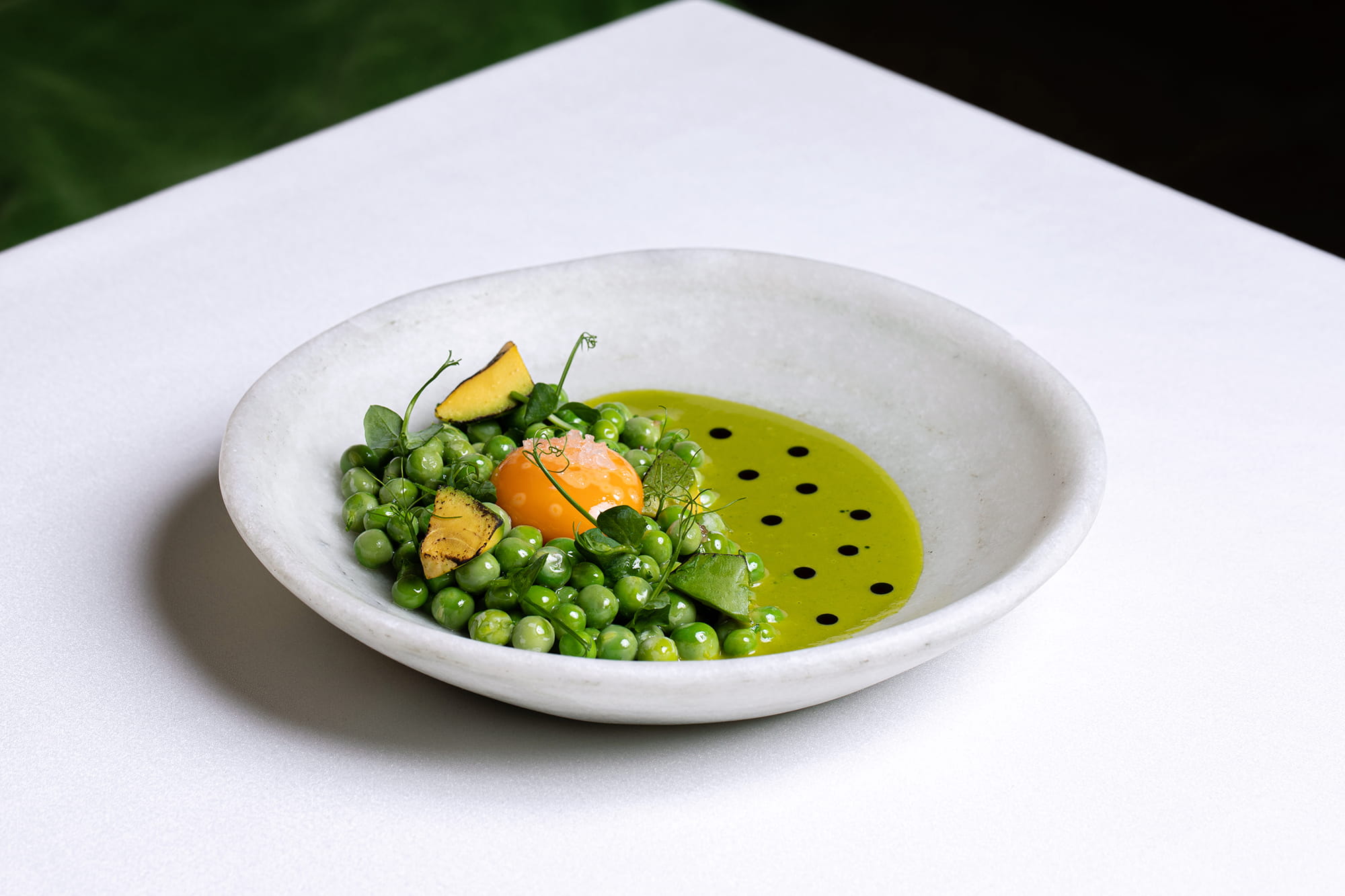 Fotografía gastronómica de plato de guisantes asados con yema de huevo, aguacate y crema verde en vajilla contemporánea