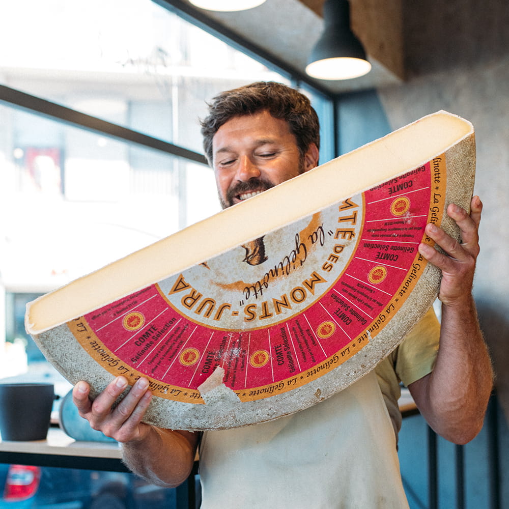Fotografía gastronómica de maestro quesero sosteniendo gran pieza de queso Comté en interior de quesería artesanal