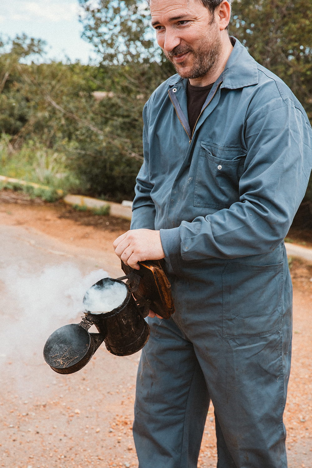 Apicultor con ahumador en mano preparando intervención en colmena durante proceso de apicultura en entorno rural