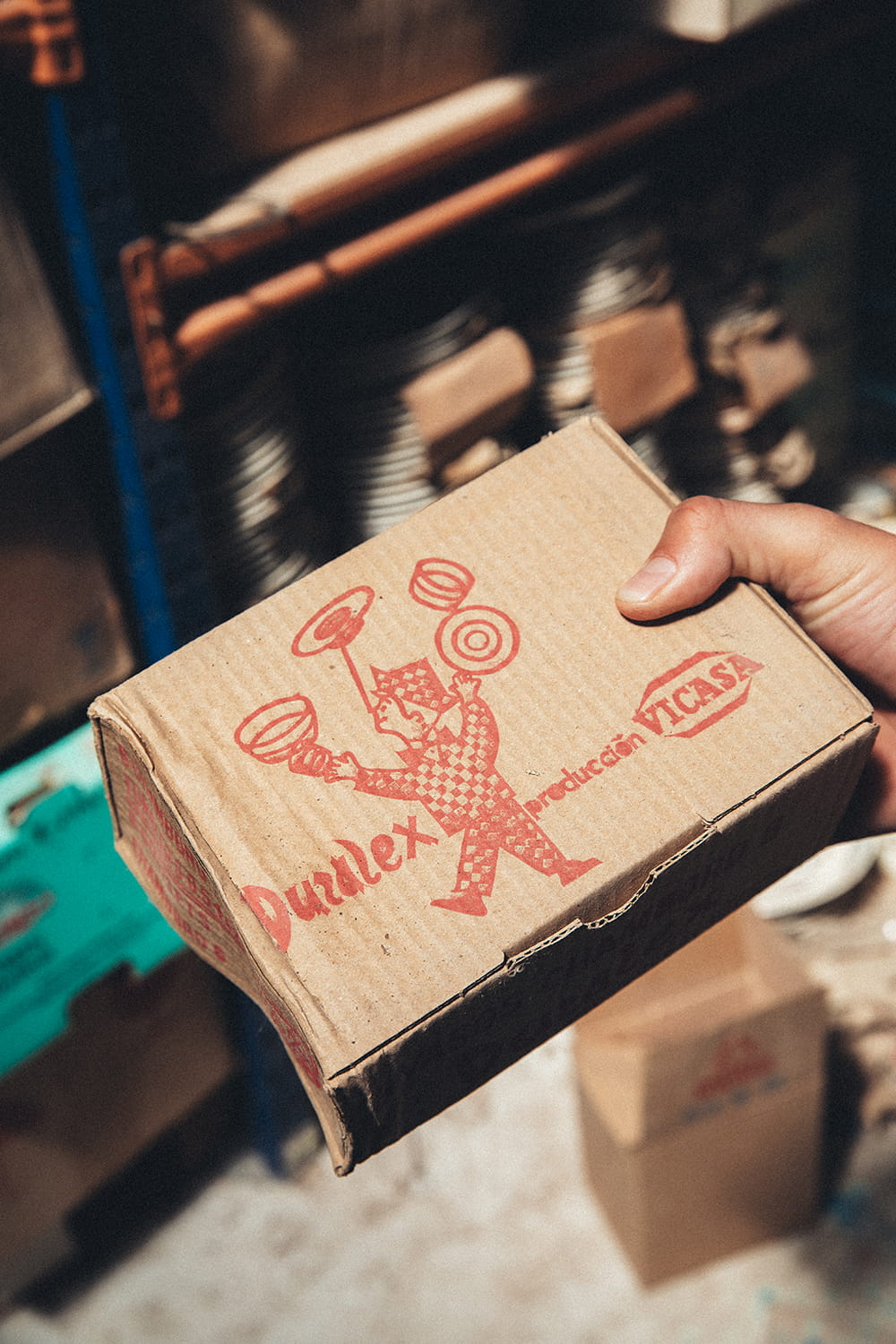 Mano sosteniendo una caja original de Duralex Producción VICASA con ilustración de malabarista en tinta roja, encontrada en la nave de Cristal Antiguo.