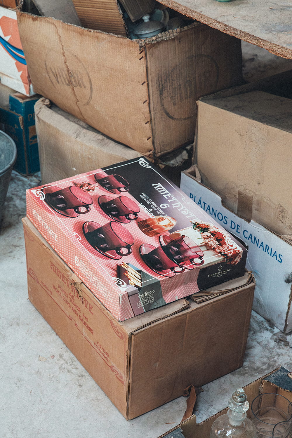 Caja original de juego de tazas de café "Amethyste" de la marca Vereco en color lila, sobre cajas de cartón antiguas en el suelo polvoriento del almacén de Cristal Antiguo.