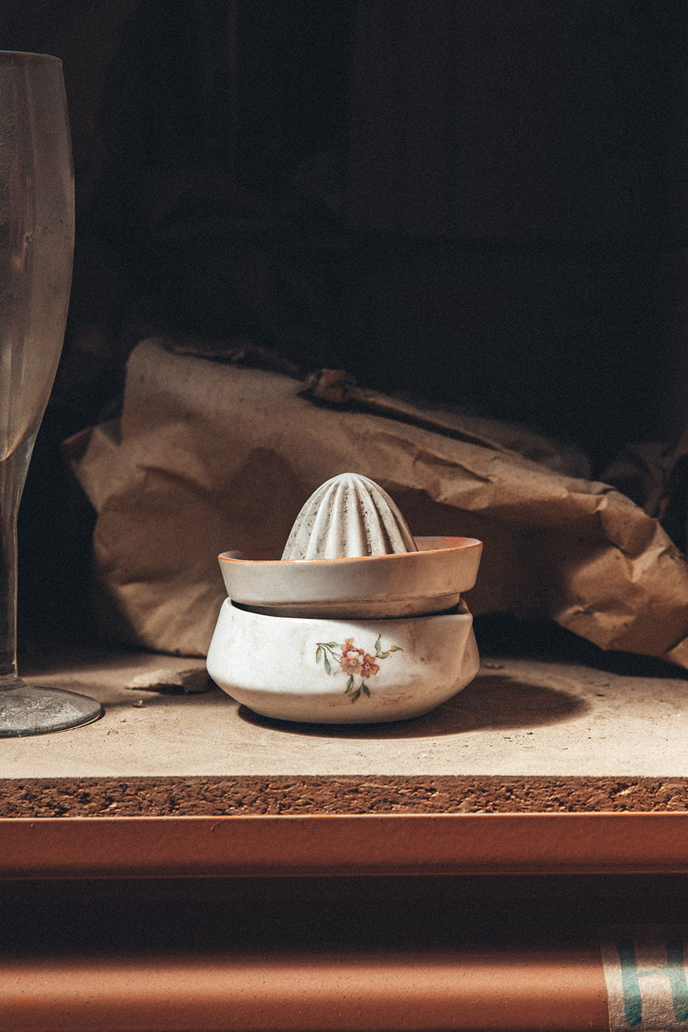 Primer plano de un exprimidor de cerámica antiguo con detalle de flores pintadas, sobre una estantería con polvo en el almacén de Cristal Antiguo