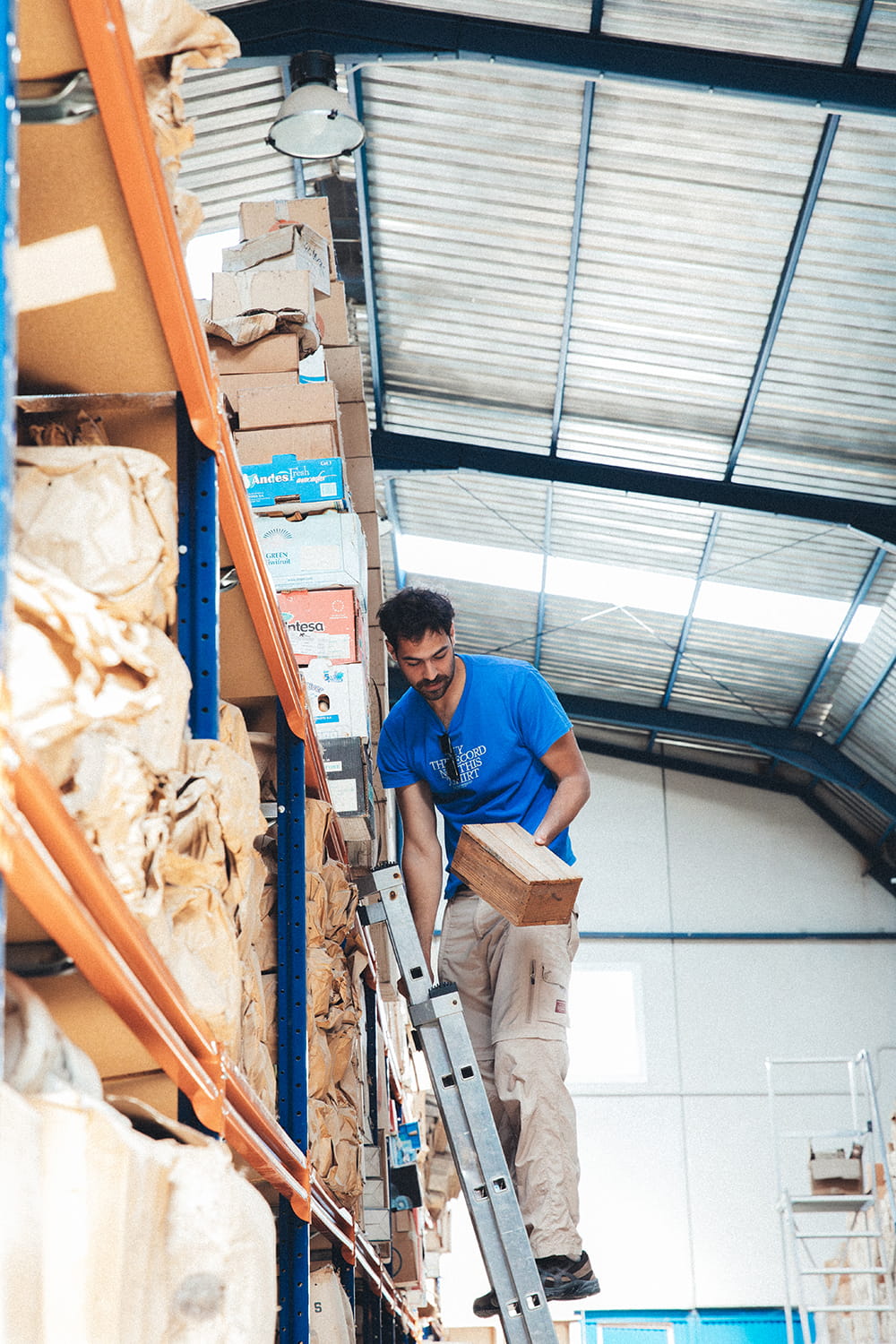Juan Bohajar subido a una escalera, cogiendo una caja de madera antigua de China de una estantería alta en el almacén de Cristal Antiguo