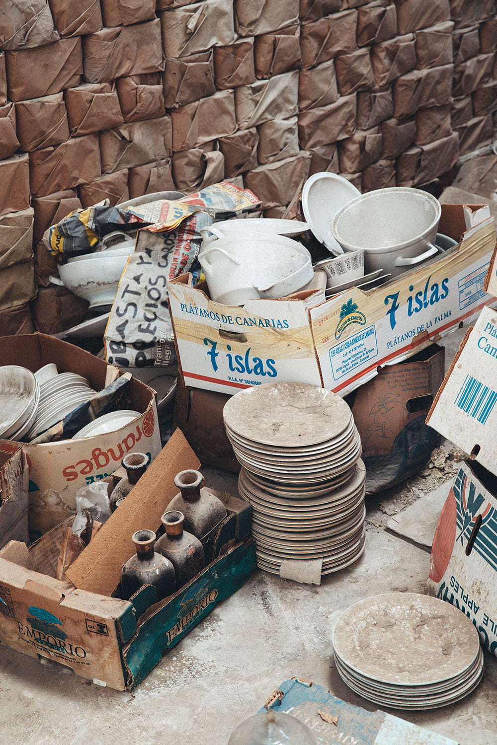 Pilas de platos polvorientos, fuentes de loza blanca y botellas antiguas de cerámica almacenadas en cajas de cartón de fruta (Plátanos de Canarias 7 Islas) sobre el suelo de la nave de Cristal Antiguo.