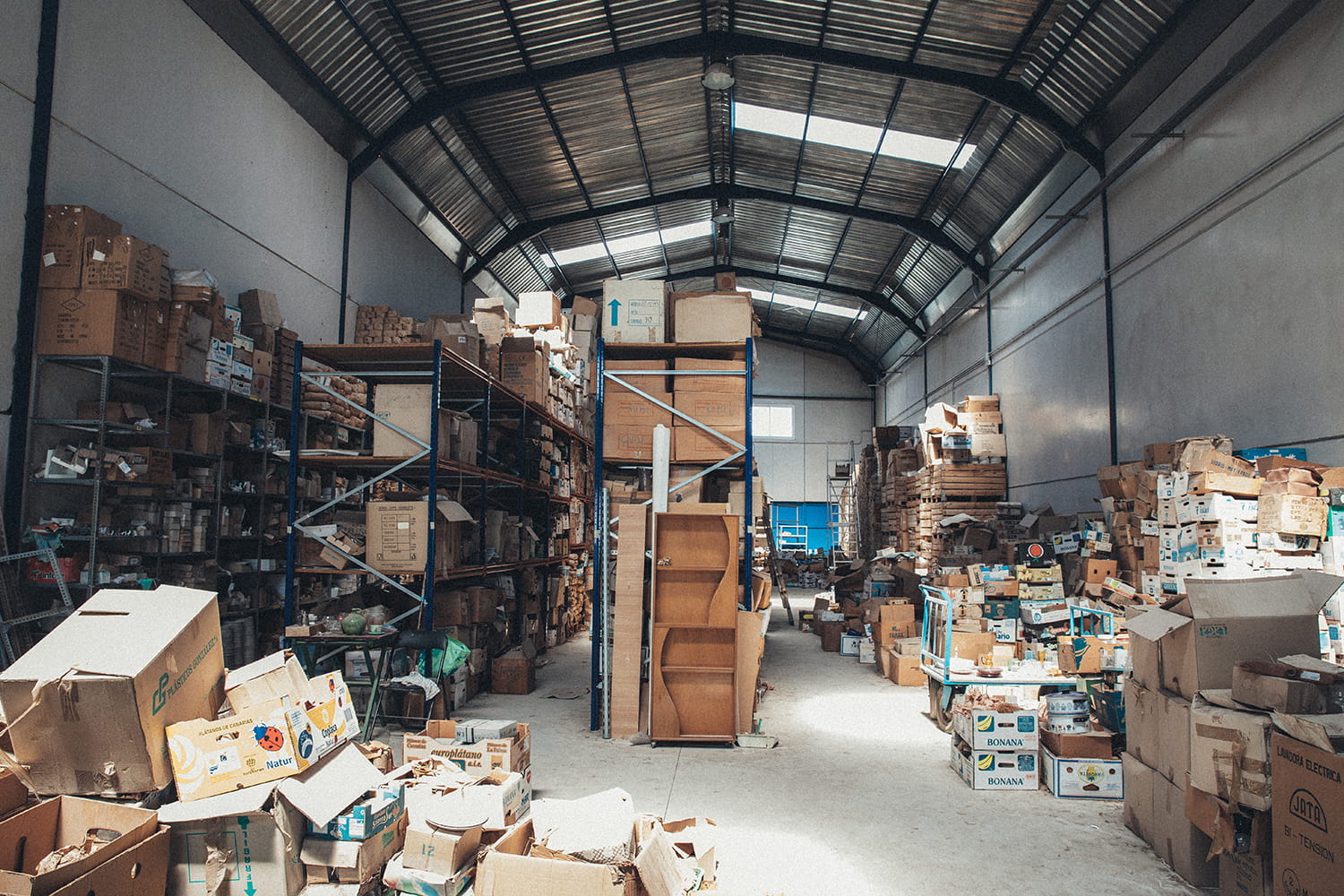 Vista general del interior de la nave industrial de Cristal Antiguo en Lorquí, con pasillos largos y estanterías repletas de cajas de cartón y madera bajo la luz natural de los tragaluces