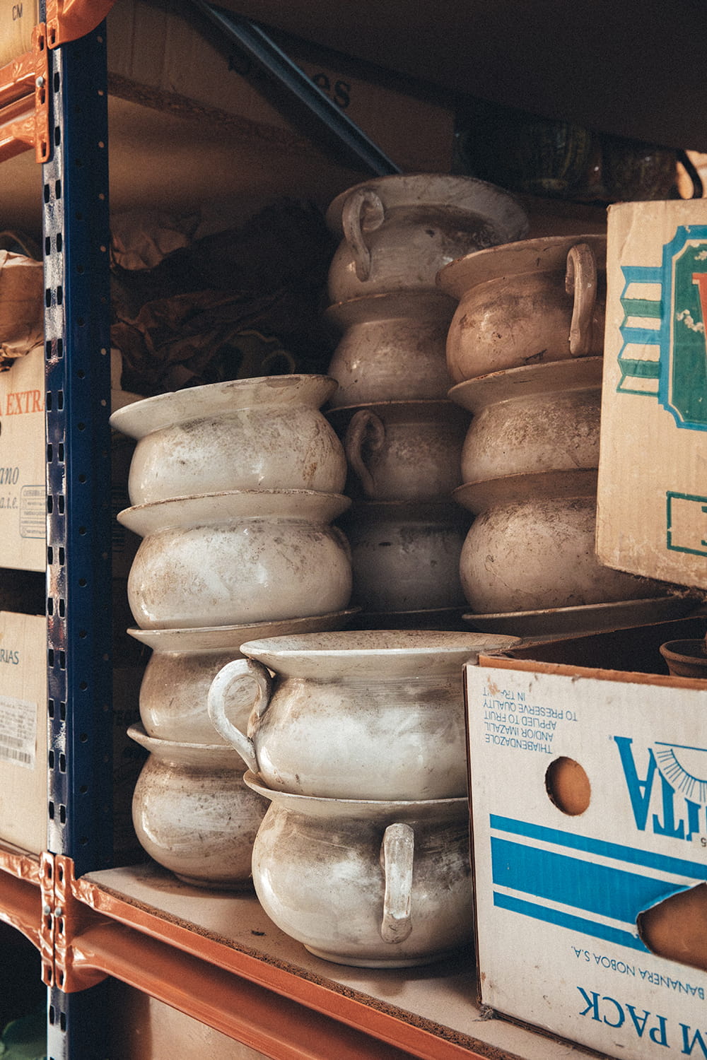 Pilas de orinales de loza blanca polvorientos almacenados en una estantería metálica del almacén de Cristal Antiguo en Lorquí, junto a cajas de cartón