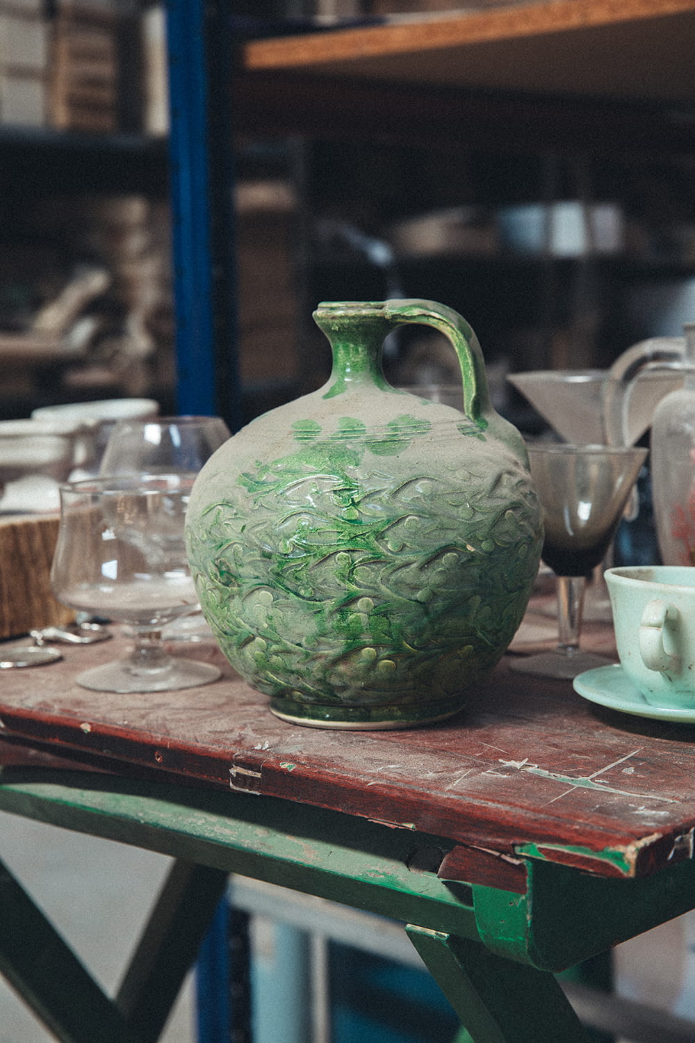 Primer plano de una orza de cerámica verde antigua con polvo, sobre una mesa de madera verde en el almacén de Cristal Antiguo