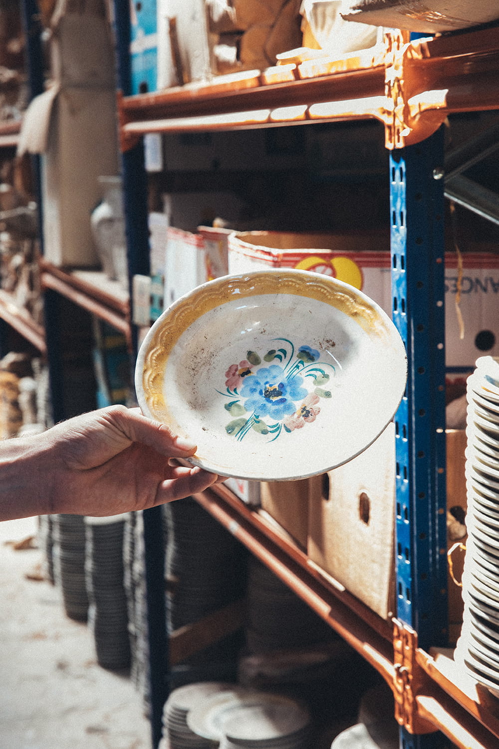 Primer plano de una mano sosteniendo un plato hondo de cerámica antigua con motivos florales pintados a mano en azul y rosa, perteneciente a la colección histórica de Cristal Antiguo.