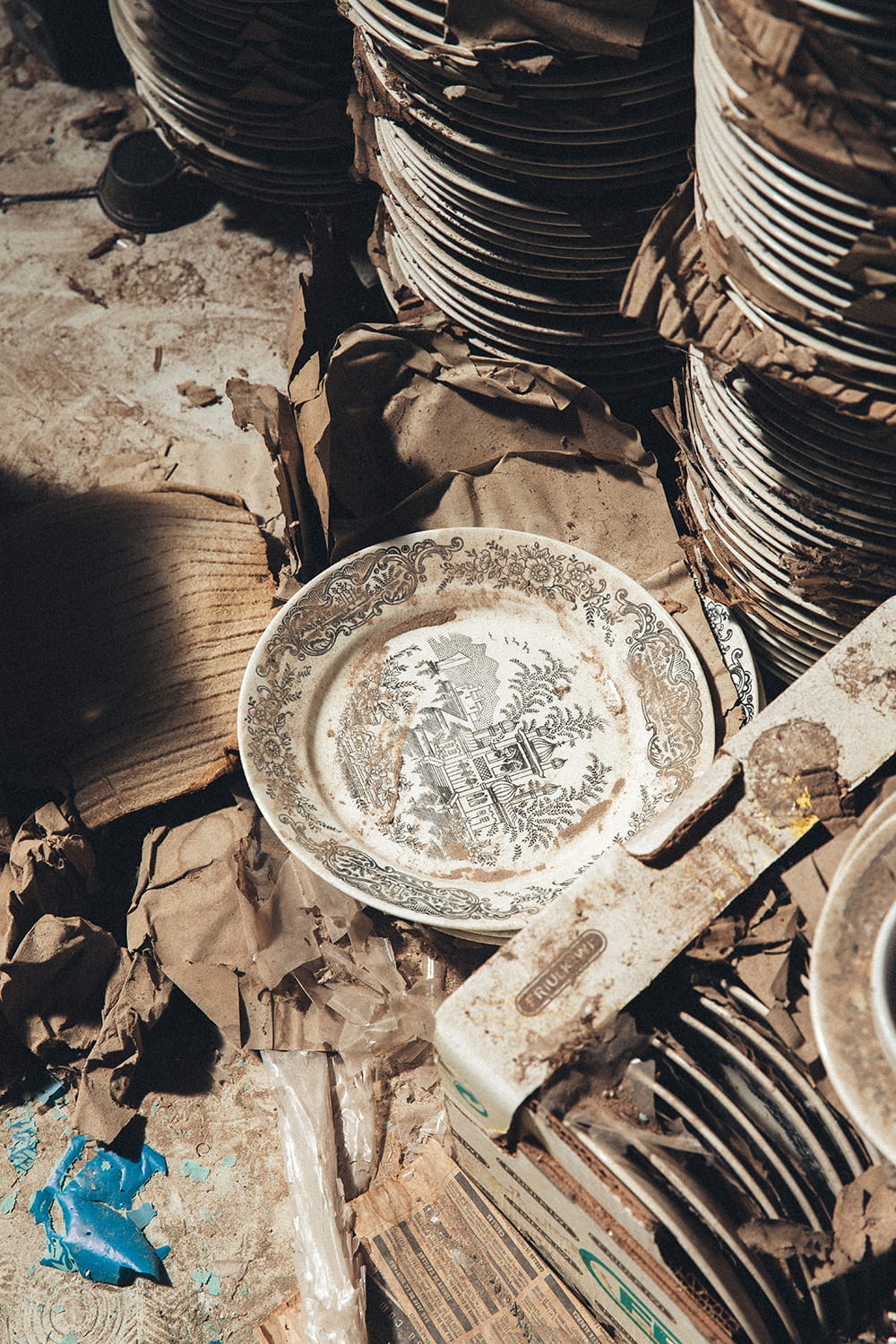 Pilas de platos de loza antigua con estampación de paisajes, rodeados de papel de embalaje original en el almacén de Cristal Antiguo